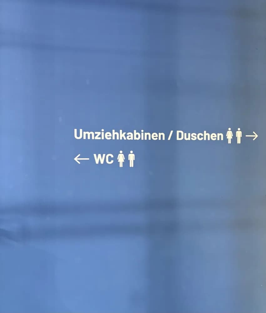Wegweiser an einer blauen Wand: „Umziehkabinen / Duschen" mit Pfeil nach rechts, „WC" mit Pfeil nach links