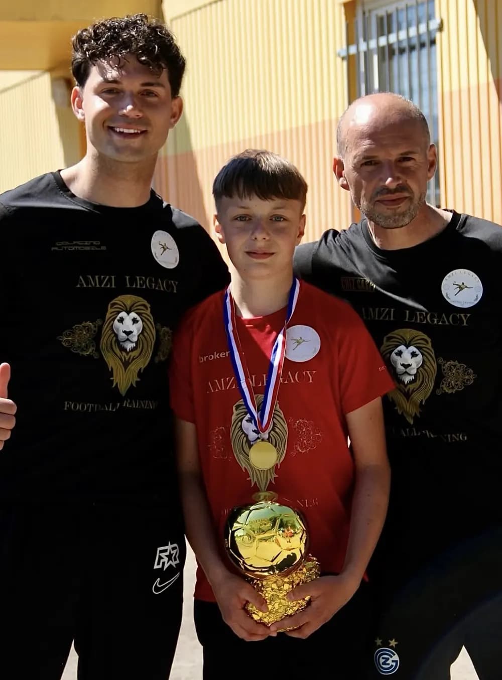 Ein Junge mit Medaille hält einen Pokal, daneben zwei Trainer