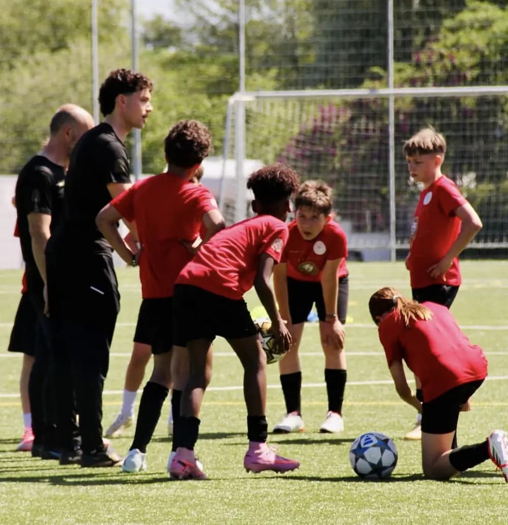 Kinder in roten Trikots üben mit dem Ball auf dem Rasen, ein Trainer steht am Rand