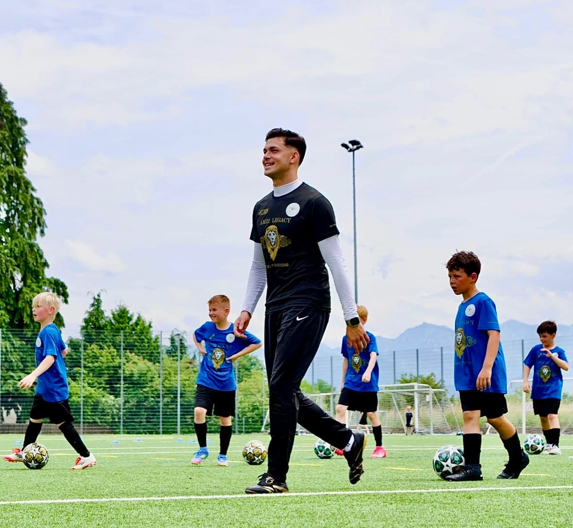 Trainer auf dem Rasenplatz, um ihn herum Kinder in blauen Amzi-Legacy-Shirts, jedes mit einem Ball