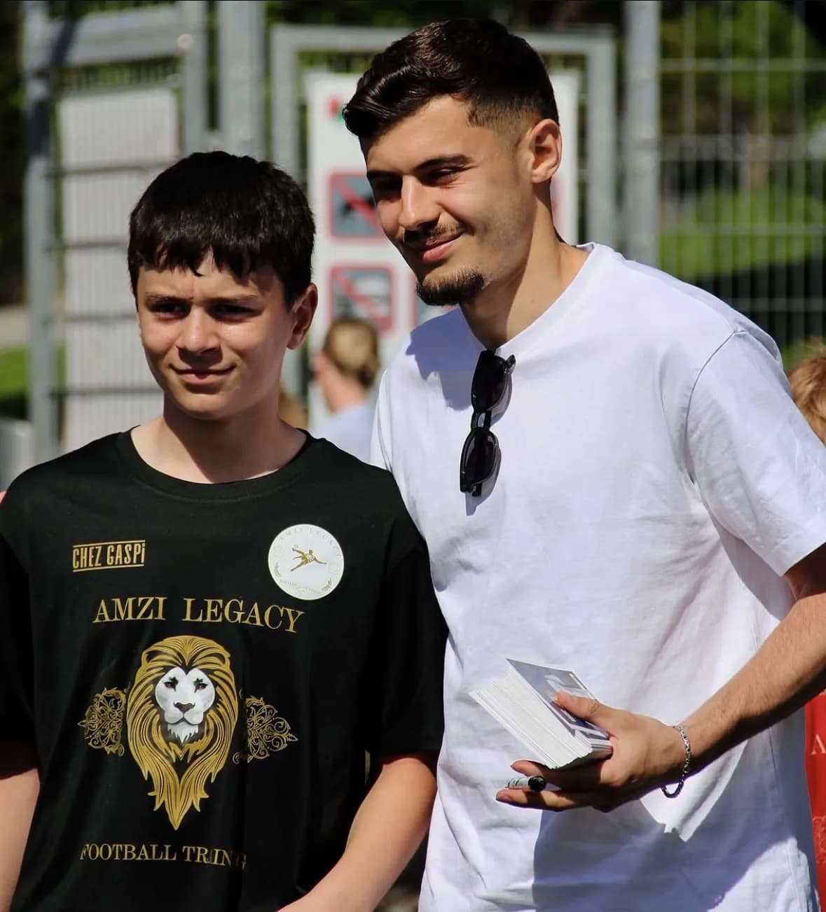 Ein Profispieler steht neben einem Jungen im schwarzen Trikot von Amzi Legacy und hält Autogrammkarten in der Hand