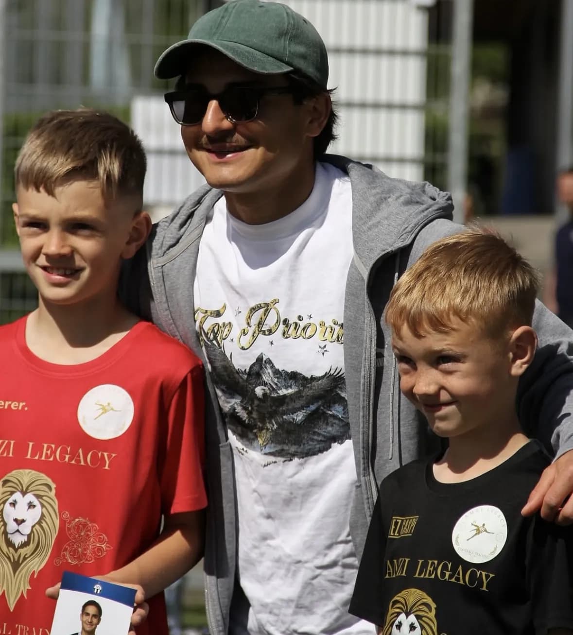 Ein Profispieler mit Kappe und Sonnenbrille steht zwischen zwei Jungen in Trikots von Amzi Legacy