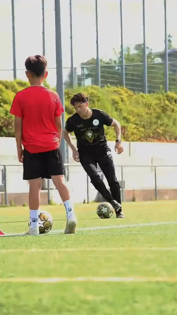 Ein Trainer führt den Ball auf einen Jungen im roten Shirt zu, auf dem Rasenplatz im Camp